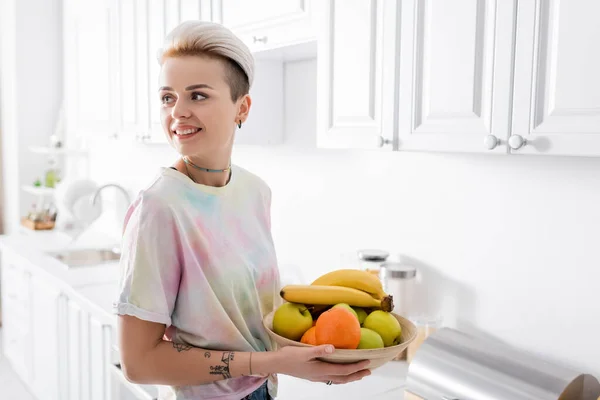 Pleased tattooed woman holding bowl with ripe fruits in kitchen and looking away — стоковое фото