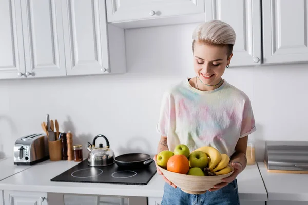 Cheerful woman with trendy hairstyle holding bowl of ripe fruits in kitchen - foto de stock