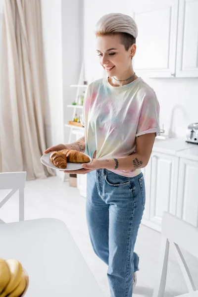 Smiling tattooed woman in jeans holding plate with delicious croissants in kitchen — Fotografia de Stock