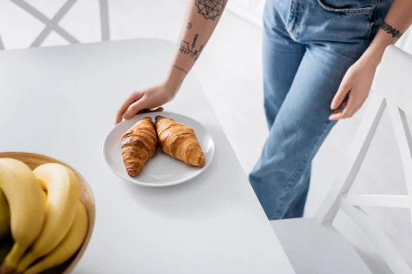 Visão parcial de mulher tatuada em jeans perto de croissants saborosos e bananas desfocadas — Fotografia de Stock