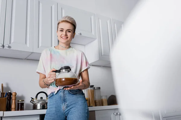 Visão de baixo ângulo da mulher feliz com pote de café olhando para a câmera na cozinha em primeiro plano borrado — Fotografia de Stock