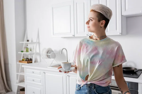 Mujer con peinado de moda sosteniendo la taza de café y mirando hacia otro lado en la cocina - foto de stock