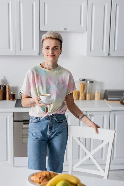 Pleased woman with cup of coffee looking at camera near blurred croissants - foto de stock