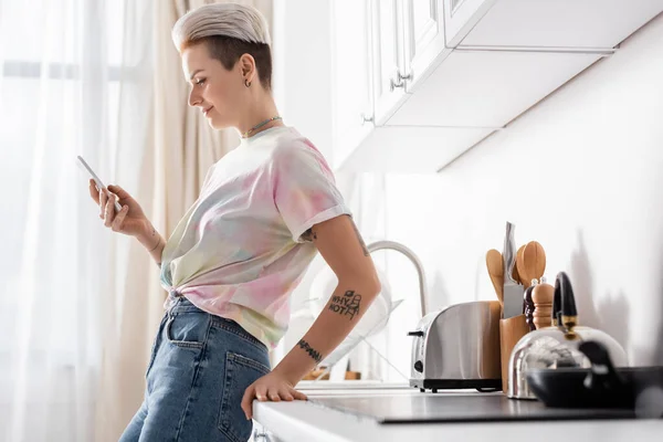 Side view of positive tattooed woman standing in kitchen with mobile phone — Stock Photo