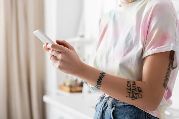 Частичный вид молодой татуированной женщины, отправляющей сообщения по мобильному телефону на кухне — стоковое фото