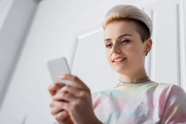 Vista a basso angolo di donna sorridente con acconciatura alla moda messaggistica su smartphone sfocato — Foto stock