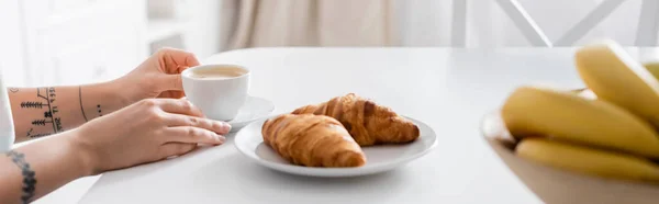 Vista parcial de mujer tatuada con taza de café cerca de croissants y plátanos borrosos, pancarta - foto de stock