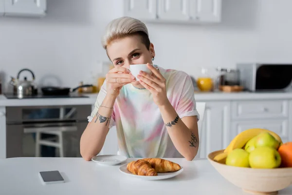 Trendy woman with coffee cup looking at camera near croissants, smartphone and fresh fruits — Foto stock