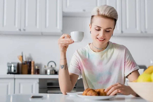 Tätowierte lächelnde Frau trinkt Kaffee mit Croissants in der Küche — Stockfoto