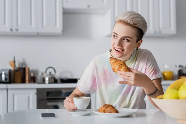 Mulher alegre segurando xícara de café e comer croissant saboroso perto do smartphone com tela em branco — Fotografia de Stock