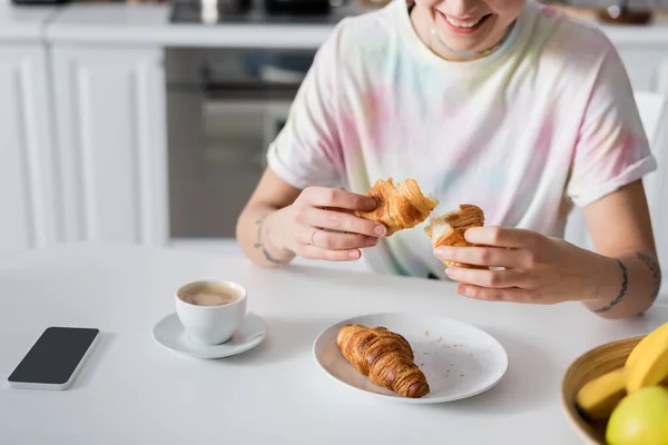 Обрізаний вид усміхненої жінки, що тримає круасан біля чашки кави та смартфона з порожнім екраном — стокове фото