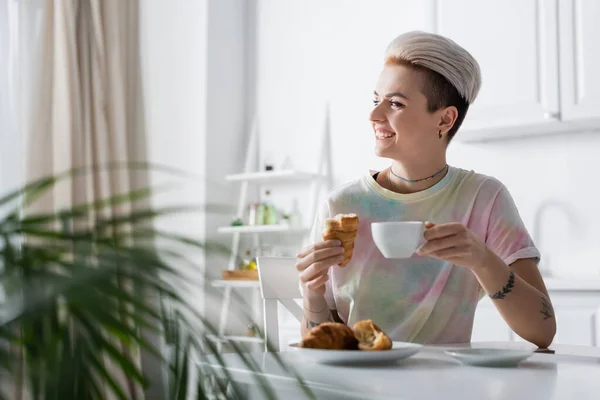 Mulher alegre com café e croissant delicioso olhando para longe na cozinha — Fotografia de Stock