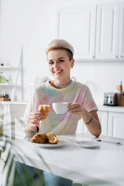 Fröhliche Frau mit Kaffeetasse und leckerem Croissant blickt in Küche in die Kamera — Stockfoto