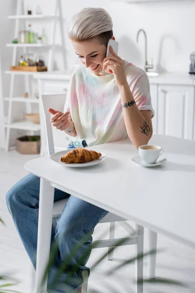 Gioiosa donna che parla su smartphone vicino a deliziosi croissant e tazza di caffè — Foto stock