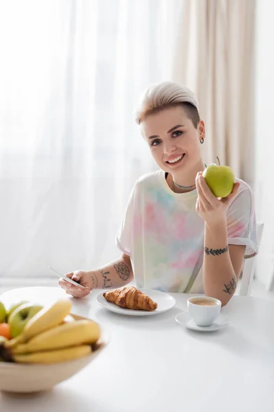 Mujer feliz y elegante sosteniendo manzana y teléfono móvil cerca de la placa con croissant - foto de stock