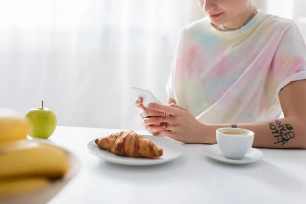 Partial view of young woman chatting on smartphone near coffee, croissant and apple — Stock Photo