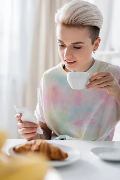 Улыбающаяся женщина с чашкой кофе с помощью смартфона возле круассана на размытом переднем плане — стоковое фото