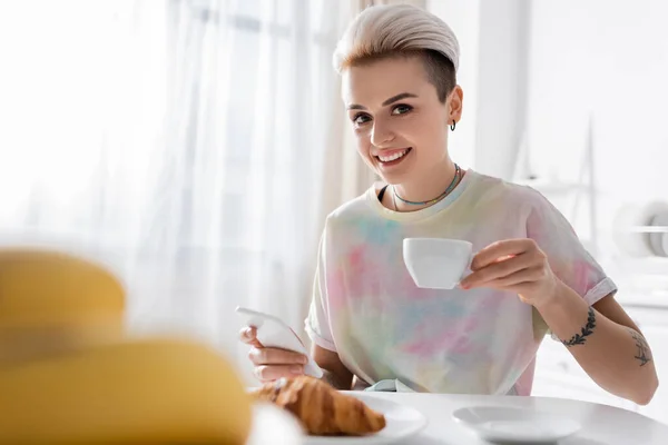 Mujer con estilo con teléfono inteligente y taza de café sonriendo a la cámara en primer plano borrosa - foto de stock