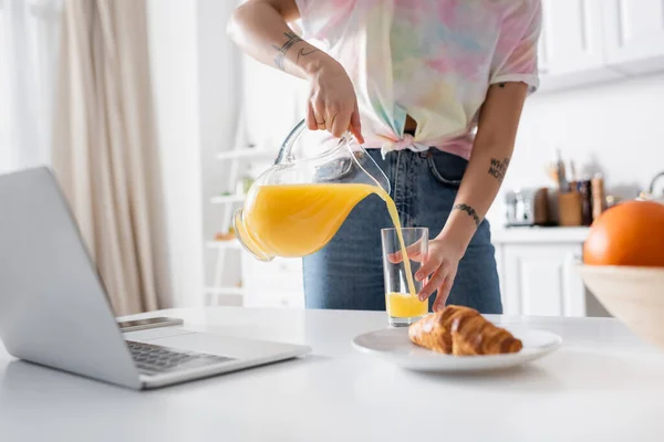 Vista cortada de mulher tatuada derramando suco de laranja perto croissant e laptop borrado — Fotografia de Stock