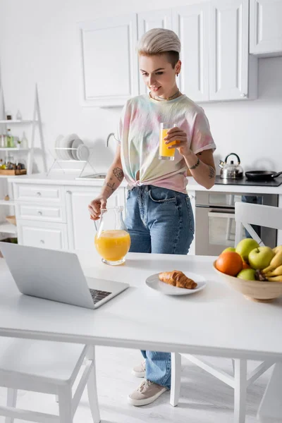 Young woman with orange juice near laptop, croissant and fresh fruits on kitchen table — Stock Photo