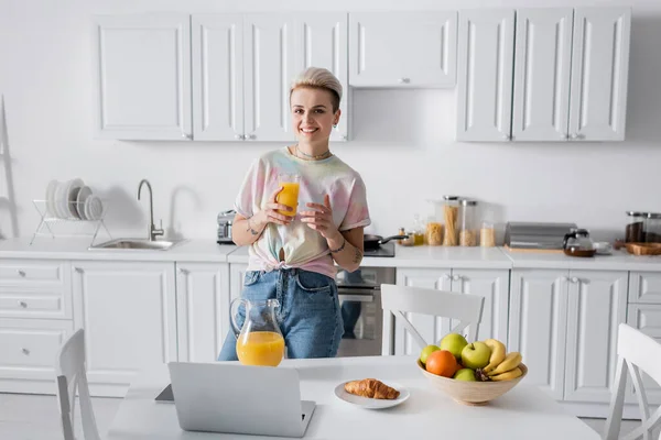 Femme à la mode avec du jus d'orange souriant à la caméra près de la table de cuisine avec ordinateur portable et petit déjeuner — Photo de stock