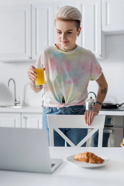 Positive woman holding glass of orange juice near blurred laptop and croissant — Fotografia de Stock