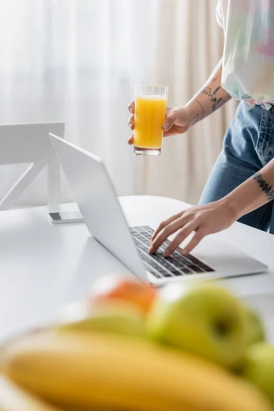Частковий вид татуйованої жінки з апельсиновим соком, що друкується на ноутбуці поблизу розмитих фруктів — стокове фото