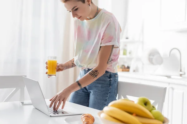 Молодая женщина с апельсиновым соком с помощью ноутбука рядом с круассаном и размытые фрукты на кухне — стоковое фото