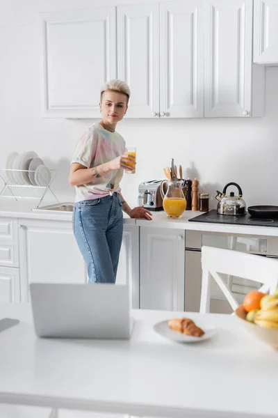 Trendy woman with orange juice looking at blurred laptop on kitchen table — Foto stock
