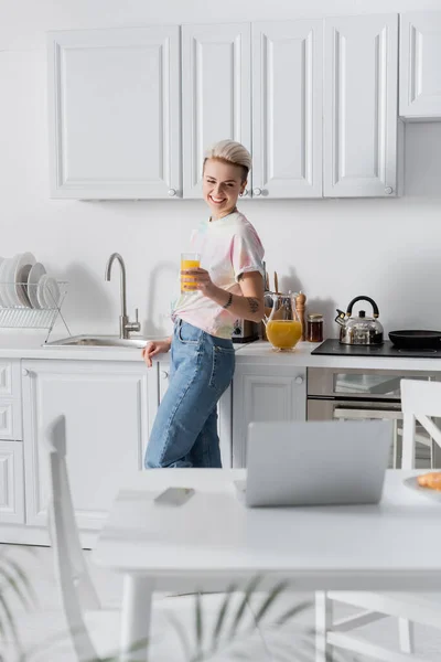 Cheerful woman with orange juice looking at blurred computer on kitchen table — Stockfoto