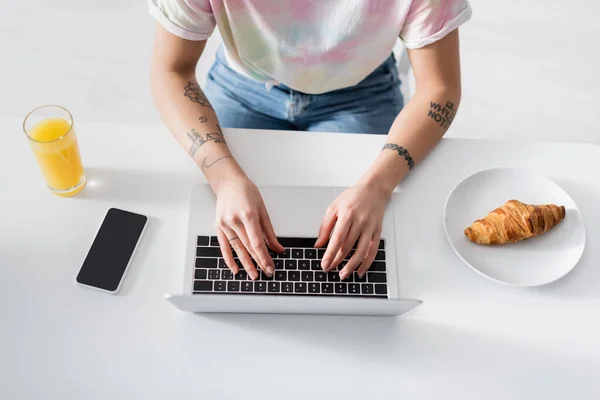 Vista superior de la mujer recortada escribiendo en el ordenador portátil cerca de jugo de naranja, croissant y teléfono móvil - foto de stock