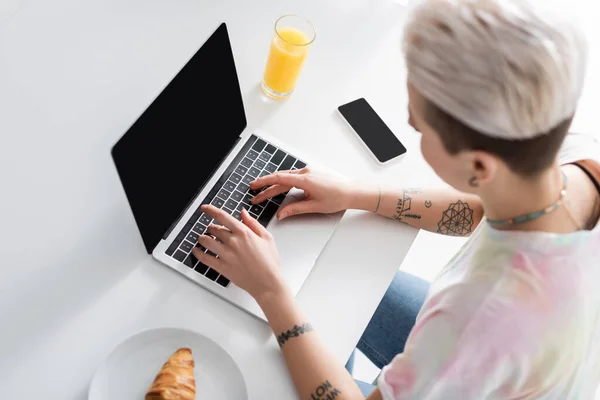 Overhead view of blurred woman typing on laptop near smartphone, orange juice and croissant — Stock Photo