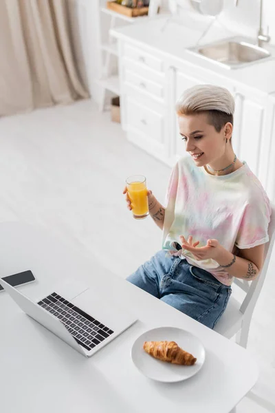 Mulher sorridente com suco de laranja apontando para laptop perto de croissant e smartphone com tela em branco — Fotografia de Stock