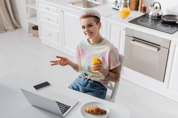Vista de ángulo alto de la mujer feliz con jugo de naranja cerca de la computadora portátil, teléfono inteligente y croissant - foto de stock