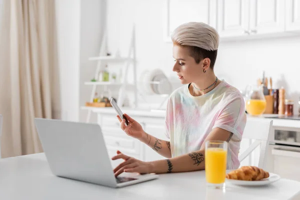 Trendy tattooed woman with mobile phone typing on laptop near orange juice and croissant — Fotografia de Stock