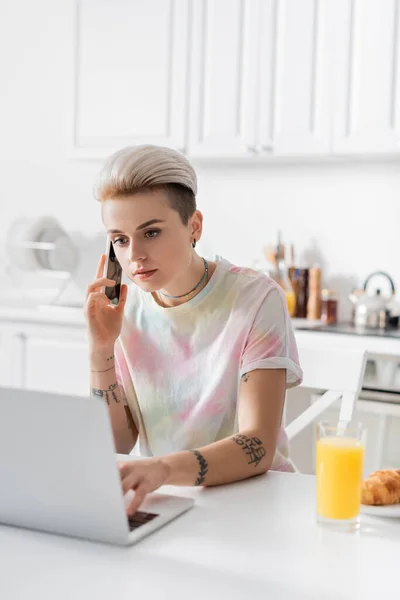 Stylish woman talking on smartphone near laptop and blurred glass of orange juice in kitchen — стоковое фото