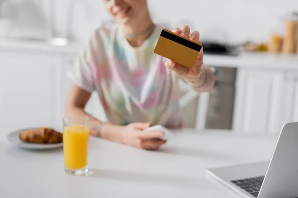 Обрізаний вид розмитої жінки з кредитною карткою біля ноутбука, склянки апельсинового соку і круасанів — стокове фото