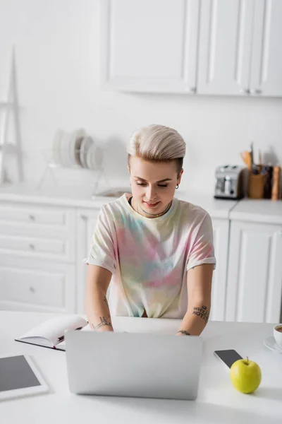 Щаслива жінка працює на ноутбуці на кухні біля смартфона, цифрового планшета та яблука — стокове фото