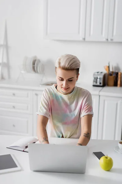 Happy woman working on laptop in kitchen near smartphone, digital tablet and apple — Stock Photo