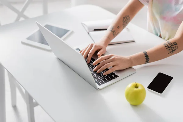 Partial view of tattooed woman typing on laptop near smartphone, digital tablet and apple — Stock Photo