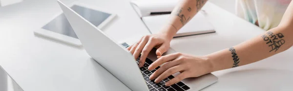 Partial view of tattooed woman typing on laptop near notebook and blurred laptop, banner — стоковое фото