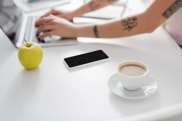 Cropped view of blurred woman typing on laptop near smartphone, coffee cup and fresh apple — Fotografia de Stock