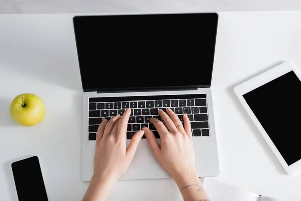 Вид зверху на жінку, що друкує на ноутбуці біля смартфона, цифрового планшета та свіжого яблука — стокове фото