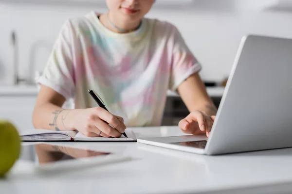 Partial view of blurred woman writing in notebook near laptop and digital tablet — Stock Photo