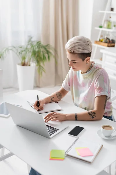 Mulher elegante escrevendo em notebook perto de gadgets e xícara de café na mesa da cozinha — Fotografia de Stock