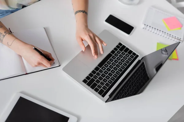 Partial view of tattooed woman writing in notebook near laptop, smartphone and digital tablet — Foto stock