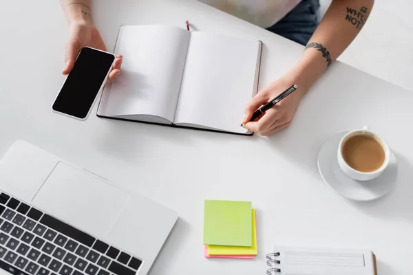 Top view of cropped woman writing in empty notebook near laptop, smartphone and coffee cup — Stock Photo