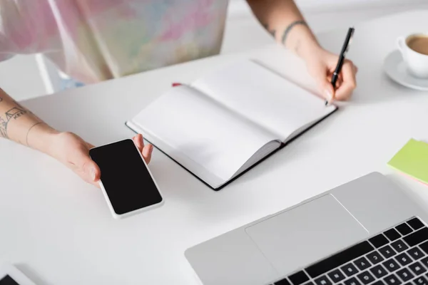 Partial view of woman with smartphone writing in empty notebook near laptop - foto de stock