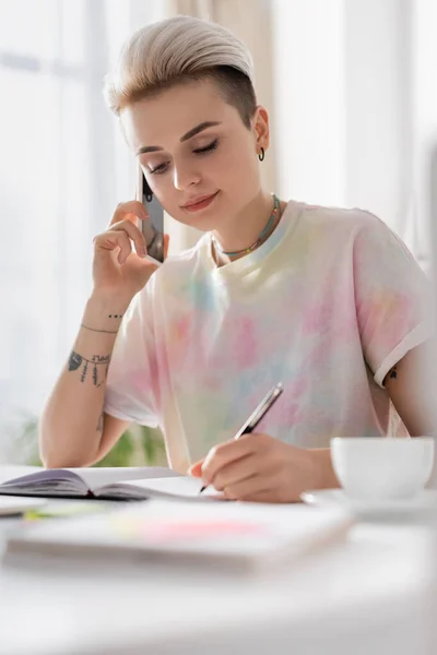 Femme élégante écrivant dans un carnet et parlant sur un téléphone mobile près d'une tasse de café floue — Photo de stock