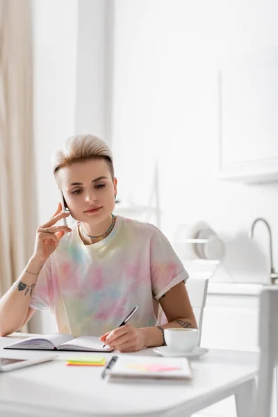 Young tattooed woman talking on smartphone and writing in notebook near coffee cup — Stockfoto